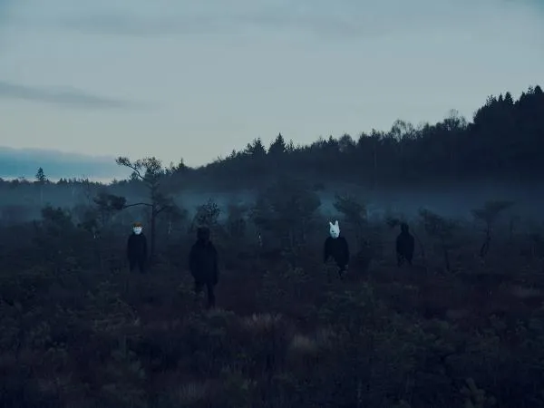 Band photo. The four band members stand in a mist covered mire wearing black clothes and animal masks, resembling from left to right a cat, a bear, a rabbit and a crow.